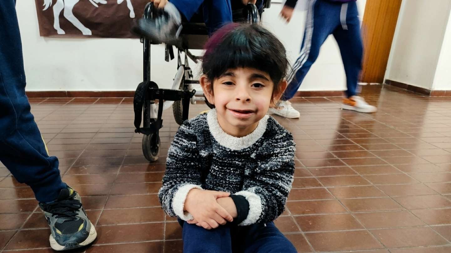 Niña sonriente sentada en el piso del instituto, con una silla de ruedas de fondo.