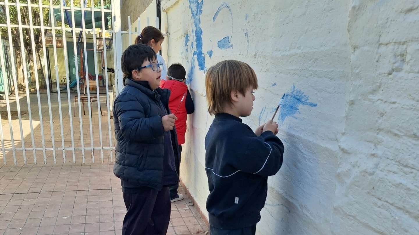 Niños pintando con pinceles y témpera azul un mural sobre una pared de ladrillos blanqueados, en el patio.